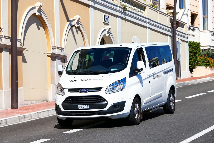 A white Ford 12-passenger van labeled 'MOBI BUS' is driving on a street with a beige and white building in the background. The van has a license plate with the number 'F216' and a wheelchair accessibility symbol on the front. The building has arched windows and a sign that reads 'EFG.' The street is lined with a sidewalk, and a lamppost is visible on the left side of the image.